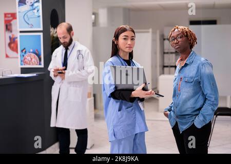 Asiatische Krankenschwester, junge afroamerikanische Frau Porträt im Krankenhaus Wartezimmer. Arzt, der der weiblichen Verwandten Medikamente des Patienten zeigt. Gesundheitsfachkraft Kontrollprogramm auf dem Smartphone in Sanatorium. Stockfoto