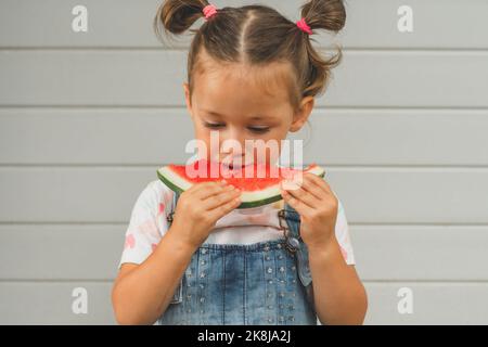 Konzentriertes kleines Mädchen 3-4 Jahre alt mit zwei Pferdeschwänzen isst Wassermelone. Kind im Freien vor dem Hintergrund des Gebäudes hält süße gesunde Früchte in Stockfoto