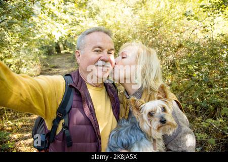 Selfie eines reifen kaukasischen Paares, das sich beim Tragen eines kleinen Hundes im Wald küsst Stockfoto