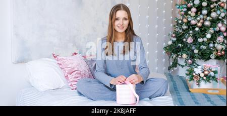 Glückliches Mädchen in blauen Pyjamas mit Geschenk in der Hand sitzt im Schlafzimmer auf Hintergrund Weihnachtsbaum. Festliche Stimmung, Winterferien und Feier des neuen Jahres. Glückliche kaukasische junge Frau Zeit Stockfoto