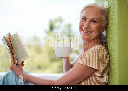 So entspannend, wie das Leben sein sollte. Eine reife Frau liest ein Buch und hat zu Hause Kaffee auf ihrem Balkon. Stockfoto