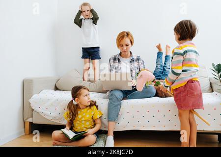 Ein Haufen Kinder spielen um ihre Mutter herum, die auf der Couch sitzt und an ihrem Laptop arbeitet. Jeder tut sein eigenes Ding. Stockfoto