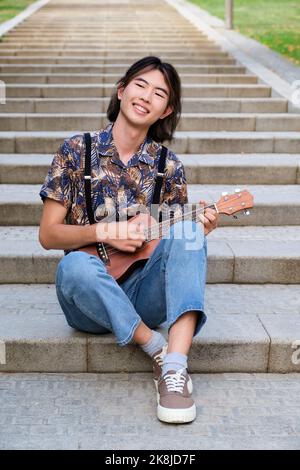 Asian Kerl lächelnd und spielt akustische Ukulele Gitarre sitzen auf Treppen. Stockfoto