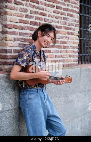 Asiatischer Kerl lächelt und spielt akustische Ukulele-Gitarre. Stockfoto