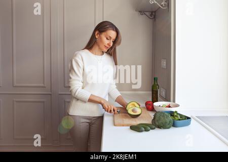 Frau kocht vegetarischen Salat mit frischem Gemüse. Modellschneiden mit Messer-Avocados, Brokkoli, Grünkochsalat, Gurken, Tomaten in der weißen Küche Stockfoto