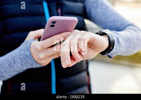 Frau mit Mobiltelefon, das an eine Smart Watch angeschlossen ist. Die Sportlerin schaut auf die Smartwatch und hält ihr Smartphone in der anderen Hand, im Freien. Fitness-FEM Stockfoto