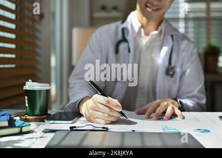 Eine kurze Aufnahme eines lächelnden Arztes in weißer Uniform, der in einem hellen Büro sitzt und eine medizinische Form ausfüllt Stockfoto