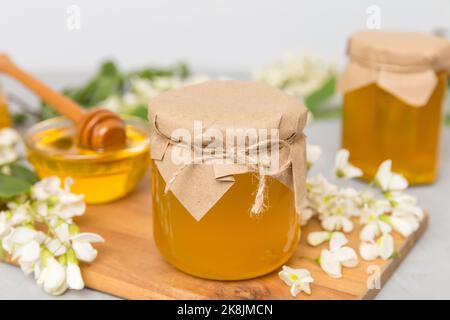 Süßes Honigglas umgab die Akazienblüten des Frühlings. Honig fließt aus einem Löffel in ein Glas. Gläser von klarem frischen Akazienhonig auf Holzgrund. Stockfoto