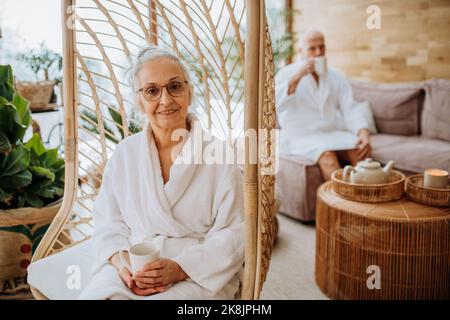 Ältere Frau, die auf einer Schaukel im Gebäude sitzt und die Kamera anschaut, ihr Mann genießt eine Tasse Tee im Hintergrund. Stockfoto