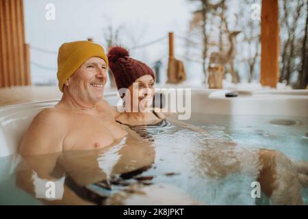 Das ältere Paar mit kitschiger Kappe genießt an kalten Wintertagen gemeinsam die Badewanne im Freien auf der Terrasse. Stockfoto