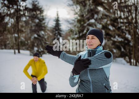 Ältere Paare wärmen sich vor der Winterwanderung im verschneiten Wald auf und strecken sich. Stockfoto