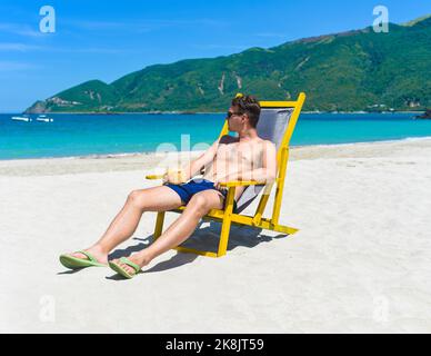 Ein junger glücklicher Mann trinkt Kokosnusssaft, der auf einem Liegestuhl an einem südchinesischen Strand in vietnam sitzt Stockfoto