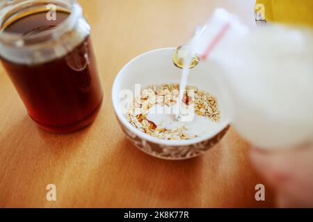 Nahaufnahme einer Frau, die Milch in eine Schüssel mit Hafer gießt. Auf dem Tisch Glas mit Honig. Stockfoto