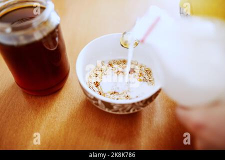 Nahaufnahme einer Frau, die Milch in eine Schüssel mit Hafer gießt. Auf dem Tisch Glas mit Honig. Stockfoto