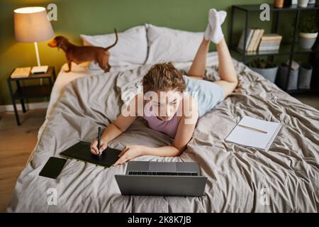 Die junge Frau, die zu Hause im Bett liegt, auf einem grafischen Tablet zeichnet, kann aus einem hohen Winkel betrachtet werden Stockfoto