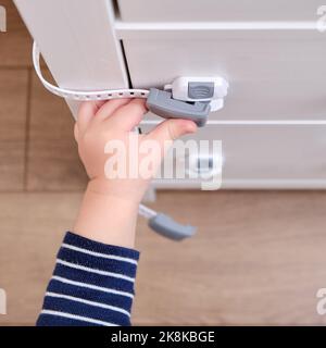 Das Baby öffnet das Kinderschloss in der geschlossenen Schublade des Schranks. Kleinkind Baby mit Hand öffnet Türschloss auf der Brust der Schubladen. Kind im Alter von einem Jahr und drei Stockfoto