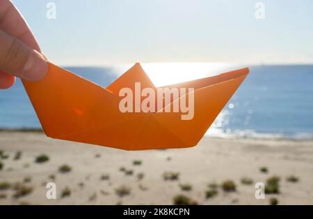 Eine Person, die mit einem orangefarbenen Papierboot auf dem Hintergrund des blauen Himmels, des Sandstrandes und des Meeres mit einem sonnigen Pfad am sonnigen Sommertag spielt. Konzept Reise Tourismus Urlaub Urlaub Traum Kindheit Spiel fantasieren Stockfoto