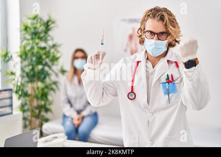 Blonder Mann in Arztuniform und medizinischer Maske mit Spritze genervt und frustriert schreiend vor Wut, schreiend verrückt vor Wut und die Hand angehoben Stockfoto