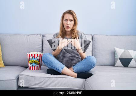 Schöne blonde Frau, die Fernsehen schaut, sitzt im Schockgesicht auf dem Sofa, sieht skeptisch und sarkastisch aus, überrascht mit offenem Mund Stockfoto