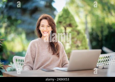 Selbstbewusste Frau mittleren Alters mit Laptop auf dem Balkon. Attraktive weibliche tragen Pullover und fröhlich lächelnd. Stockfoto