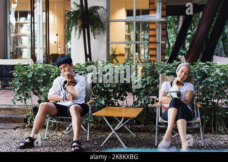 Asiatisches Paar, das einen kleinen Hund auf einem Stuhl im Garten im Freien hält. Stockfoto