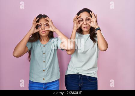 Junge Mutter und Tochter, die auf rosa Hintergrund stehen und versuchen, die Augen mit den Fingern zu öffnen, sind müde und müde für die morgendliche Müdigkeit Stockfoto