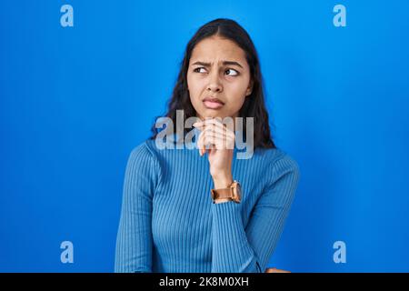 Junge brasilianische Frau, die über blauem, isolierten Hintergrund steht, denkt besorgt über eine Frage, besorgt und nervös mit der Hand am Kinn Stockfoto