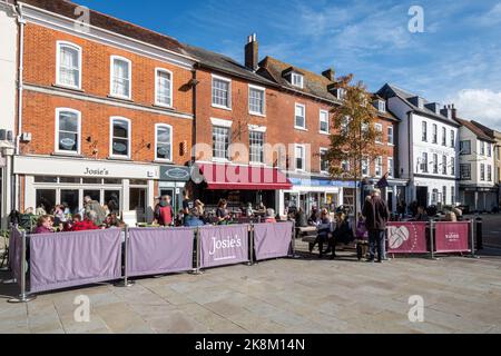 The Market Place im Stadtzentrum von Romsey, Hampshire, England, Großbritannien, mit Menschen, die an sonnigen Tagen vor Cafés und Restaurants an Tischen sitzen Stockfoto