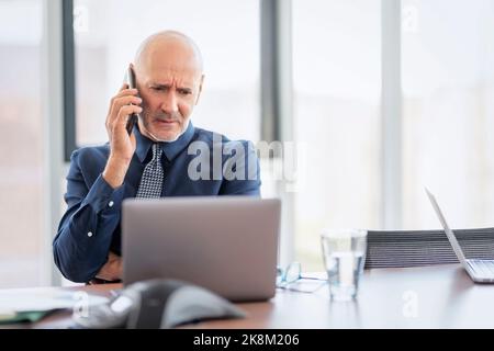 Ein besorgter Geschäftsmann, der einen Anruf tätigen musste, während er hinter seinem Laptop im Büro saß. Stockfoto