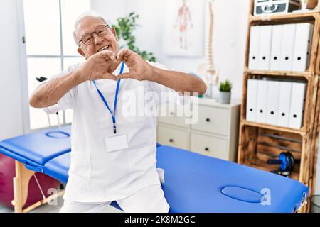 Leitender Physiotherapie-Mann, der in der Schmerzheilungsklinik arbeitet, lächelt verliebt Herz-Symbol-Form mit den Händen. Romantisches Konzept. Stockfoto