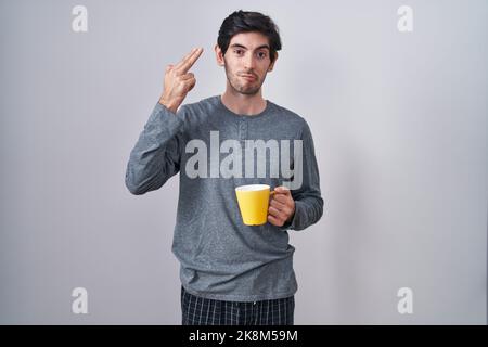 Junger hispanischer Mann mit Pyjama, der eine Tasse Kaffee trinkt und sich dabei umbringt, wie eine Pistole, eine Selbstmordgeste, Hand und Finger an den Kopf zu zeigen. Stockfoto