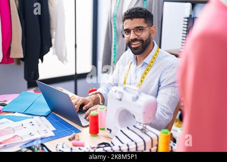 Junger arabischer Mann, der im Schneidergeschäft mit einem Laptop lächelnd zulächelt Stockfoto