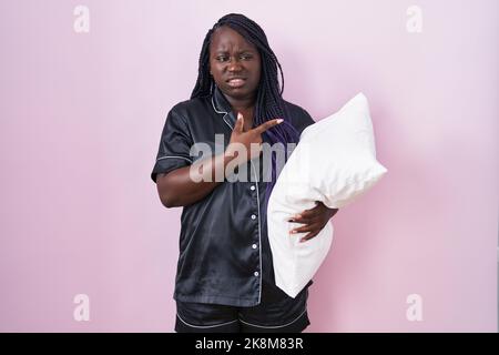 Junge afrikanische Frau trägt Pijama umarmt Kissen zeigt zur Seite besorgt und nervös mit Zeigefinger, besorgt und überrascht Ausdruck Stockfoto