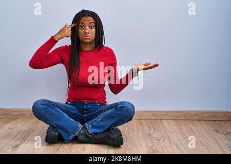 Der junge afroamerikaner mit Zöpfen, die zu Hause auf dem Boden sitzen, ist verwirrt und verärgert über die offene Handfläche, die den Kopierraum zeigt und mit dem Finger auf die Stirn zeigt Stockfoto