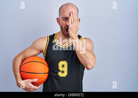 Junge Glatze Mann mit Bart trägt Basketball Uniform halten Ball gähnend müde Abdeckung halb Gesicht, Auge und Mund mit der Hand. Gesicht schmerzt vor Schmerzen. Stockfoto