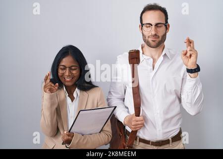 Gemischtes Geschäftspaar mit Brille gestikulend Finger überquert lächelnd mit Hoffnung und geschlossenen Augen. Glück und abergläubisches Konzept. Stockfoto