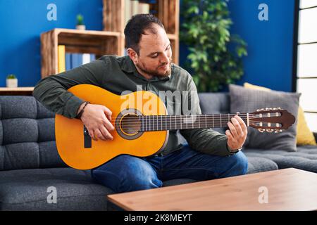 Junger hispanischer Mann spielt klassische Gitarre und sitzt zu Hause auf dem Bett Stockfoto