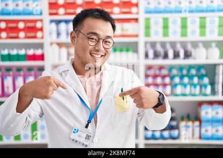 Der junge Mann aus China, der in der Apotheke arbeitet, sieht selbstbewusst mit einem Lächeln im Gesicht aus und zeigt sich mit stolzen und glücklichen Fingern. Stockfoto