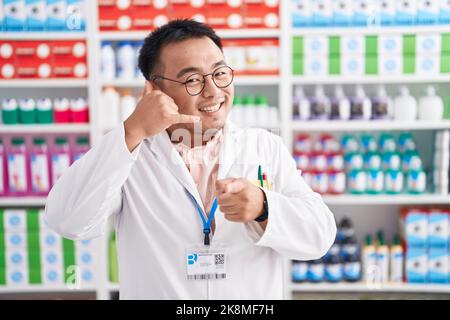 Chinesischer junger Mann, der in der Apotheke arbeitet, lächelt und telefonische Gesten macht und auf Sie zeigt. Rufen Sie mich an. Stockfoto