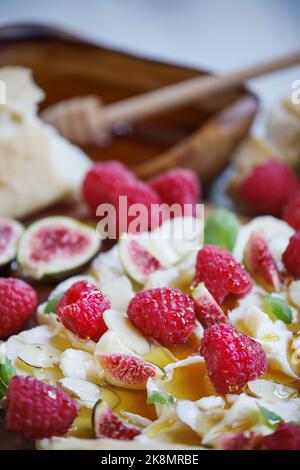 Buttertafel mit frischen Himbeeren, Feigen, in Scheiben geschnittenen Mandeln, Basilikumblättern und mit Honig beträufelt. Selektiver Fokus auf Abb. im unteren Teil des Bildes. Stockfoto