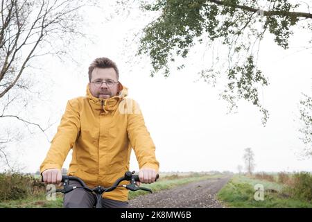 Attraktiver Mann in einem gelben Windbreaker auf einem Fahrrad, an einem nebligen Morgen, auf einer Landstraße mit Feldern im Hintergrund. Lifestyle. Stockfoto