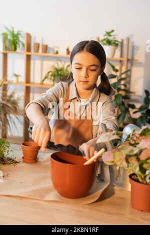 Kind in Schürze Gießen Boden in Blumentopf in der Nähe von Pflanze und Gießkanne zu Hause, Stock Bild Stockfoto