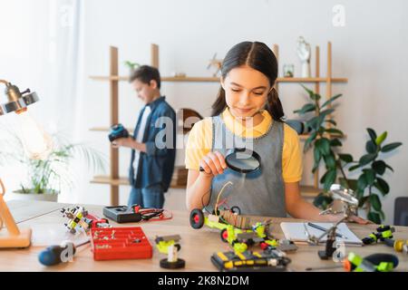 Lächelndes Kind mit Lupe in der Nähe des Robotikmodells zu Hause, Stockbild Stockfoto