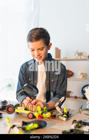 Lächelnder Junge hält Lupe in der Nähe verschwommenes Robotermodell zu Hause, Stockbild Stockfoto