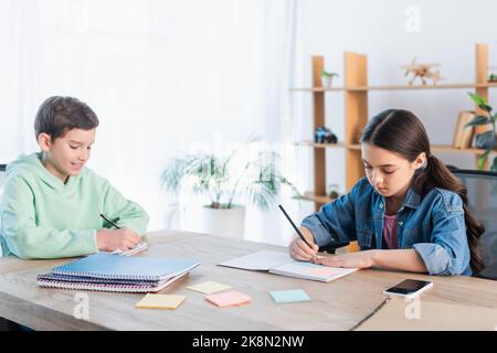 Lächelnder Junge und konzentriertes Mädchen schreiben in Notizbüchern in der Nähe von leeren Haftnotizen, Stockbild Stockfoto