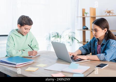 Positiver Junge, der im Notebook schreibt, und Mädchen, die auf dem Laptop tippt, während sie zu Hause studiert, Stockbild Stockfoto