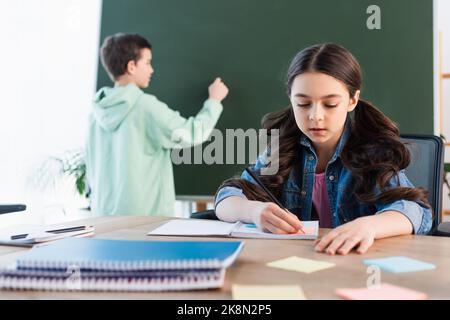 Brünette schulmädchen schreiben in Notebook in der Nähe Junge auf Kreidetafel im Hintergrund, Stock Bild Stockfoto