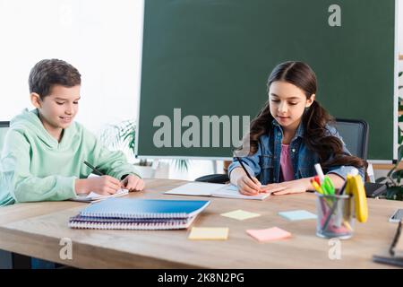 Lächelnder Junge, der in einem Notizbuch neben dem Mädchen und einer Tafel in der Schule schrieb, Stockbild Stockfoto