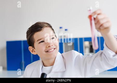 Fröhlicher Teenager Junge, der unscharfe Reagenzglas ansieht, während er im Labor chemische Experimente macht, Stockbild Stockfoto