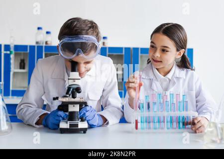 Lächelndes Mädchen hält Reagenzglas in der Nähe Freund Blick in Mikroskop während der chemischen Experiment, Stock Bild Stockfoto